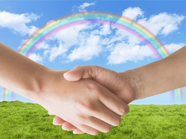 草原の向こうの青空にきれいに掛かった虹。その前で二つの手ががっちりと握手。