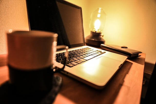 机上にノート型パソコン，コーヒーカップ，システム手帳が置いてあるランプの灯る夜のデスク