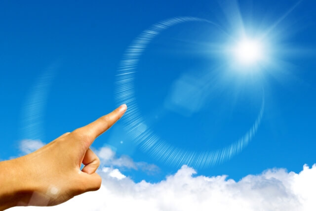 青空に輝く太陽の光球と光のリング。それを指す人差し指。