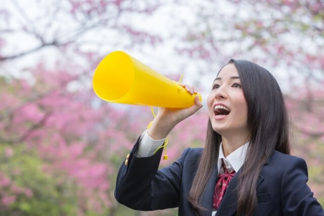 桜を背景に黄色いメガホンを翳して大きな口を開け応援しているブレザー制服の一人の女子学生。