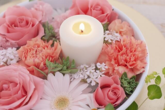容器に収まるピンク，白の種々の花々。その中に小さな炎の灯った白い１本の蝋燭。