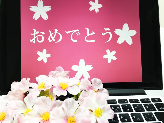 ノート型パソコンの赤い画面に映し出された白抜きの桜花と「おめでとう」の文字。キーボートの上にはピンクの花の固まり。